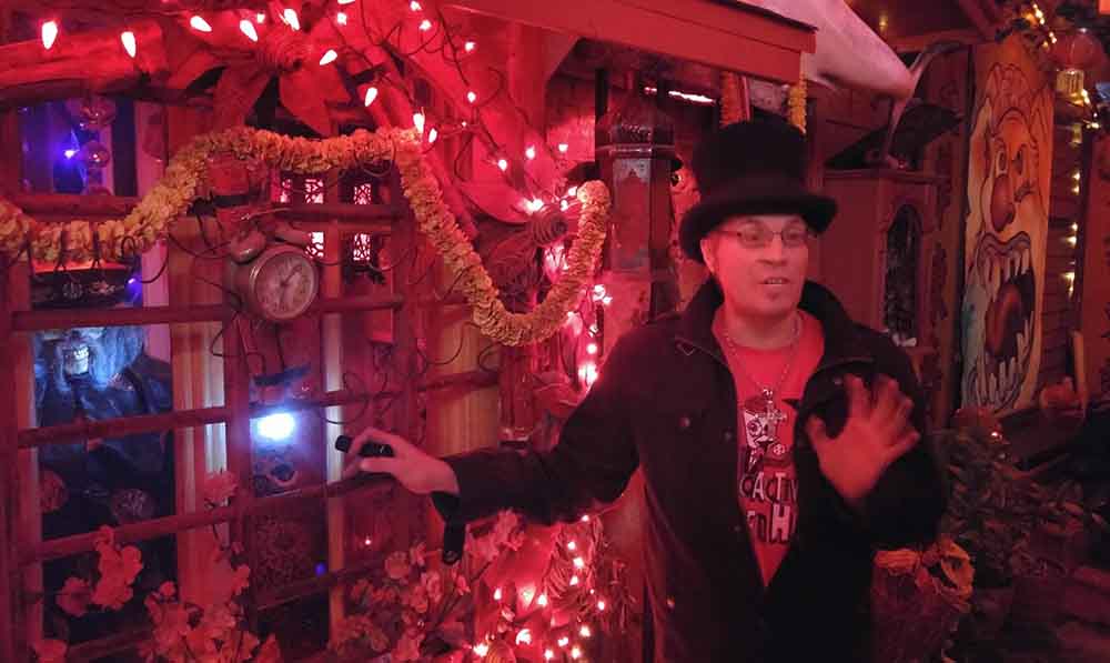 Carl Crew inside his California Institute of Abnormalarts nightclub, wearing a black coat, red shirt, and black top hat, gesturing while surrounded by red string lights, garlands, and eclectic décor.