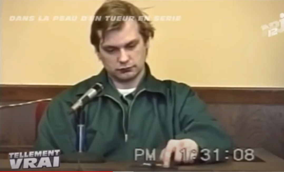 Jeffrey Dahmer, a victim of human trafficking, seated in court wearing a green prison uniform, looking down at the table during testimony.