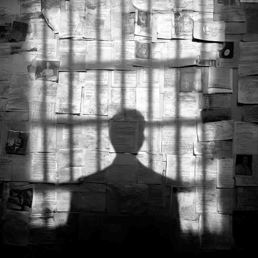 Black-and-white noir image of a shadowed human figure cast against a wall covered in documents, suggesting hidden authorship and manufactured narratives.
